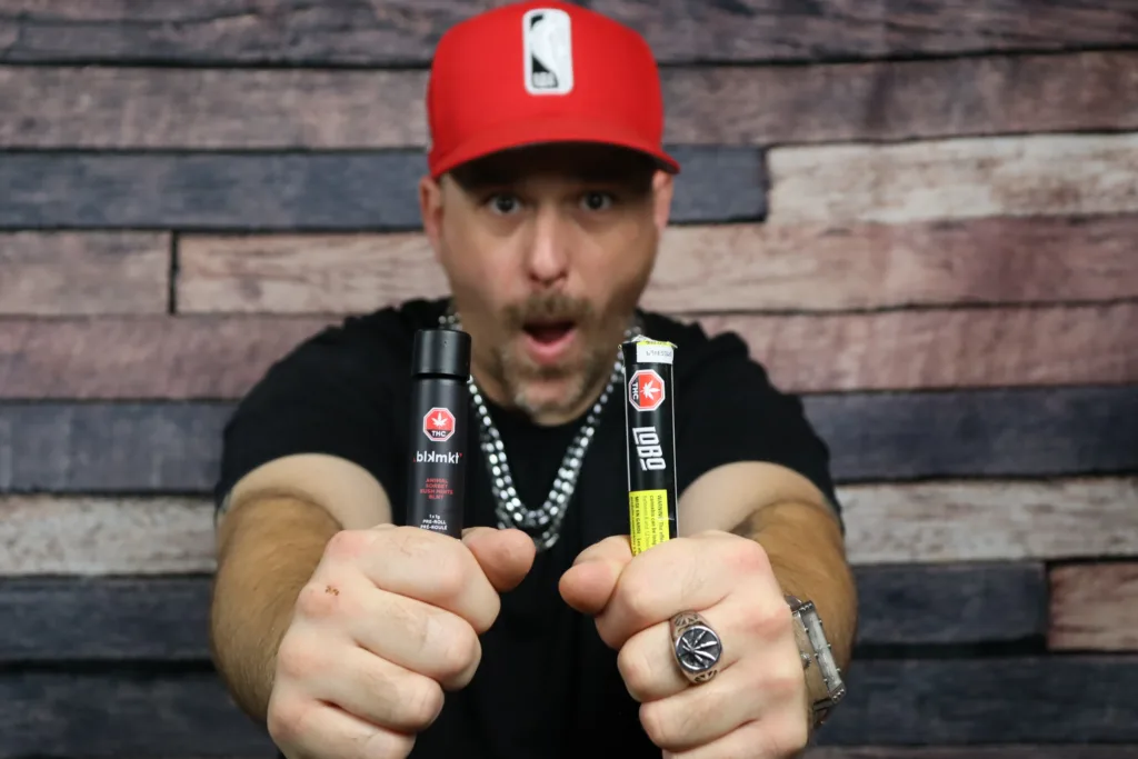 man in red hat and black shirt holding up pre rolls in front of wood background